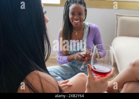 Two young women enjoy a relaxing conversation while sitting on the floor and sharing a glass of red wine The setting is intimate and cheerful, focusin Stock Photo