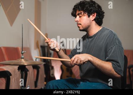 A young man with focus and passion plays the drums in a warmly lit living room He holds drumsticks expertly, engaging intensely with his cymbals Stock Photo