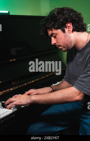 A young man with curly hair is engrossed in playing the piano, his fingers gracefully touching the keys, in a dimly lit room with a green hue enhancin Stock Photo