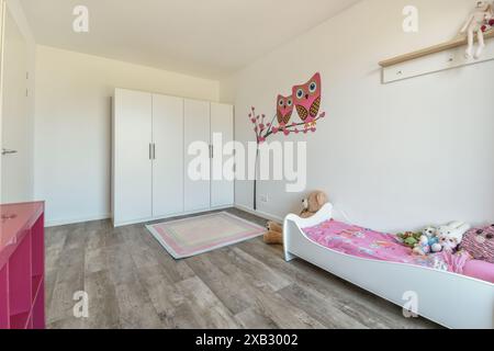 A well-lit child bedroom in Duintopstraat featuring a white bed with colorful bedding, a large wardrobe, and charming owl wall decals adding playful v Stock Photo