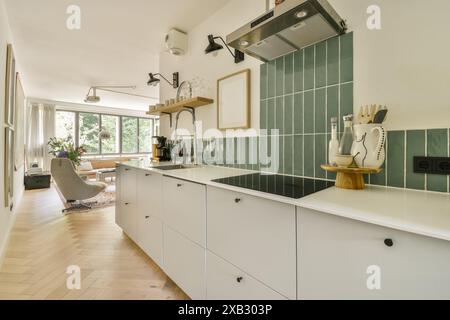 A chic modern kitchen featuring white cabinetry, green tile backsplash, and a bright living space in the background. Stock Photo