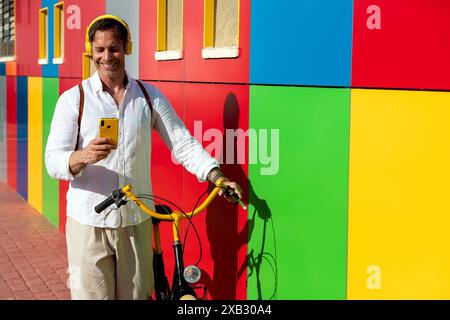 Smiling man with headphones using smartphone and leaning on a colorful wall with his yellow bike Stock Photo