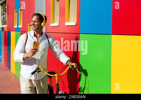A cheerful man in casual attire stands holding a yellow bike against a vibrant multicolored wall while using a smartphone and wearing headphones Stock Photo