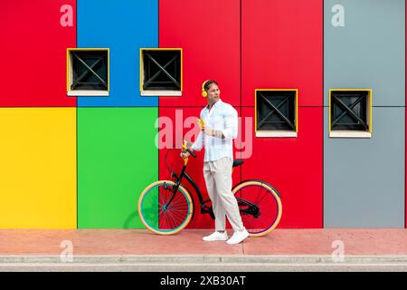 A man in casual clothing stands with a bright colored bicycle in front of a vividly painted wall with square windows Stock Photo
