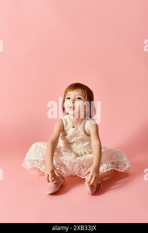 Adorable girl in white dress sitting gracefully on pink backdrop. Stock Photo
