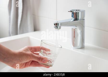 Thirsty woman's hand filling glass with fresh pure drinkable tap water from faucet in white bathroom, concept of natural clean aqua at home Stock Photo