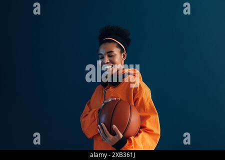 Smiling woman with a basketball wearing orange sportswear and wireless headphones. Young female with curly hair holding a basketball. Stock Photo