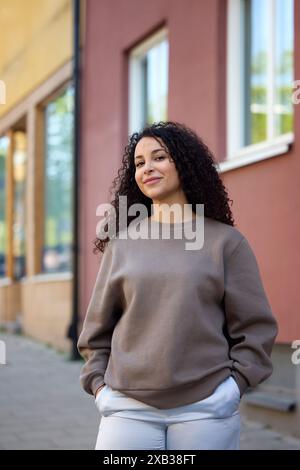 Happy curly haired woman standing with hands in pockets against building Stock Photo