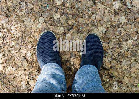 Men's legs in blue jeans and slip-on shoes, standing on the ground covered with withered leaves, first-person view Stock Photo