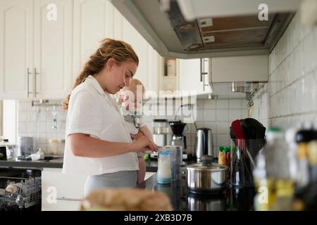 Woman carrying toddler son while preparing milk bottle at kitchen counter Stock Photo