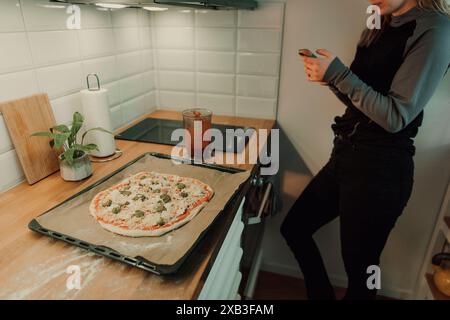 Midsection of woman using smart phone while standing by kitchen counter with homemade pizza in tray at home Stock Photo