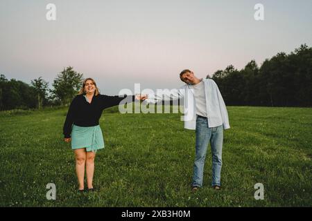 Full length portrait of happy young couple holding hands standing on grass against clear sky at sunset Stock Photo