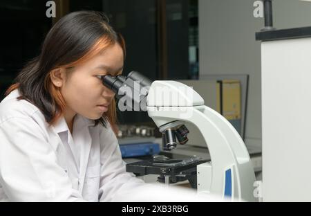 Cute scientist schoolgirl in lab coat looks through microscope for study microbiology in laboratory. Student girl child use lab equipment doing scienc Stock Photo