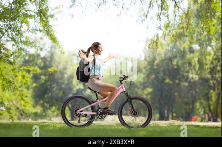 Full length profile shot of a woman with a backpack riding a bicycle and spreading arms in a park Stock Photo