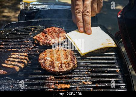 Male hand putting cheese on the top of beef patties frying on the grill, close-up view Stock Photo