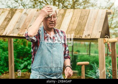 Senior man wearing a plaid shirt and overalls, wiping sweat from his forehead while working in a garden, symbolizing gardening activities and outdoor Stock Photo