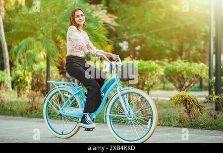 Portrait of beautiful smiling young woman riding a bicycle at sunset, looking at camera Stock Photo