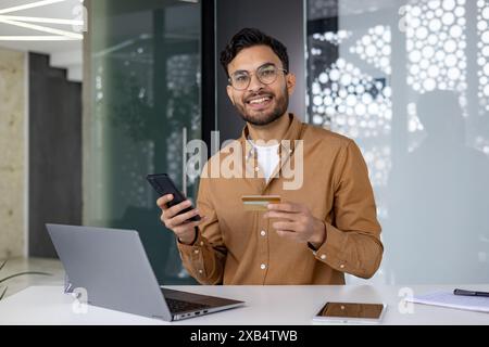 Portrait of a young Indian man sitting in a modern office at a table with a laptop, holding a mobile phone and a credit card. Stock Photo