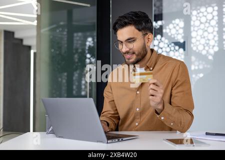 A young Indian man is sitting in an office at a desk, working on a laptop and using a credit card. Stock Photo