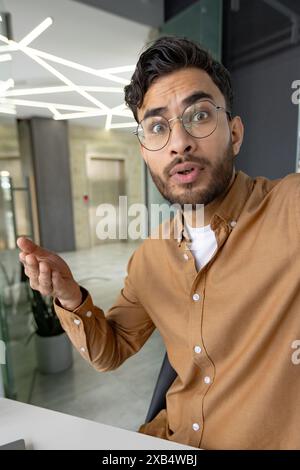 Vertical close-up photo of a worried and serious young Muslim man in a shirt and glasses sitting at a desk in the office and talking on a video call, holding a phone in his hand. Stock Photo