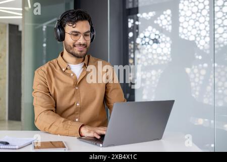 Smiling man with headphones working on a laptop in a modern office setting, focused and engaged. Stock Photo