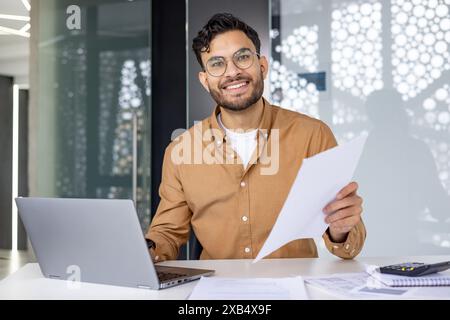 Portrait of a smiling young Muslim man working in the office with a laptop and documents, looking confidently at the camera. Stock Photo