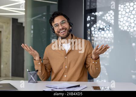 Smiling man with headset working and shrugging in modern office setting, expressing a casual and relaxed atmosphere Stock Photo