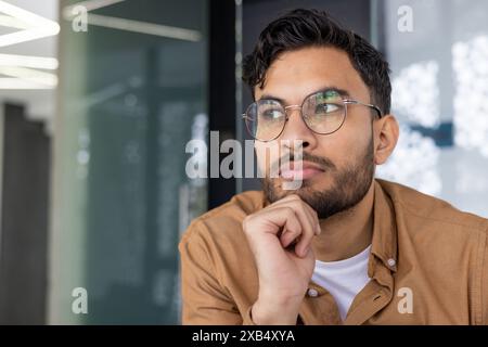 Close-up of a thoughtful young man wearing glasses and looking away, contemplating an idea while sitting indoors. Stock Photo