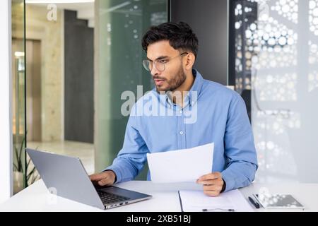 Young professional in glasses working on laptop and reviewing documents in a modern office setting. Concentration and productivity concept. Stock Photo