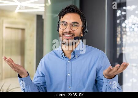 Close-up portrait of a smiling young Indian man in glasses and a blue shirt wearing a headset is in the office, looking at the camera and spreading his hands. Stock Photo