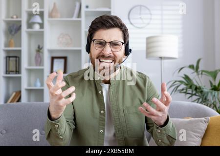 An angry young man wearing glasses and a headset speaks emotionally and angrily on a video call, gesturing with his hands to the camera. Stock Photo