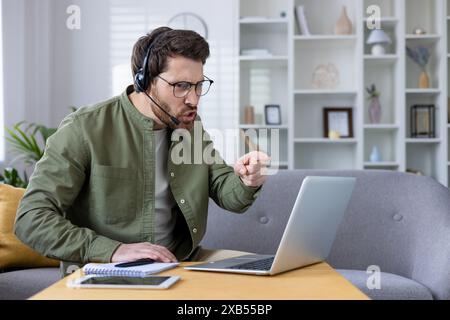 Annoyed young man in headset sitting on sofa at home and talking on video call via laptop, emotionally arguing pointing finger at monitor. Stock Photo