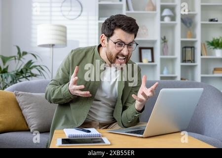 Angry young man in glasses and green shirt sitting on sofa at home in front of laptop and gesturing emotionally at screen, got bad news, talking on video call. Stock Photo