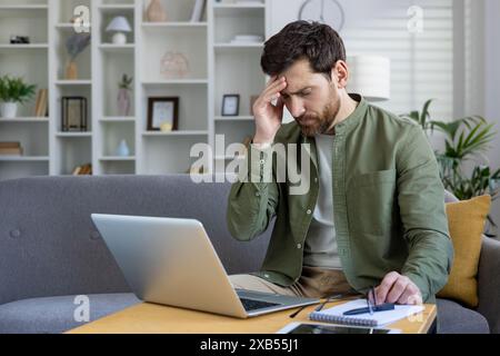 Tired young man sitting at home on sofa in front of laptop and holding hand to head, feeling severe pain and pressure, studying and working remotely. Stock Photo
