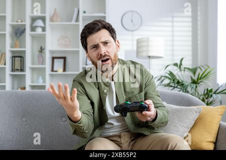 Photo of a sad young man sitting at home on the sofa with a joystick in his hands and looking disappointedly at the camera, having lost an online game. Stock Photo