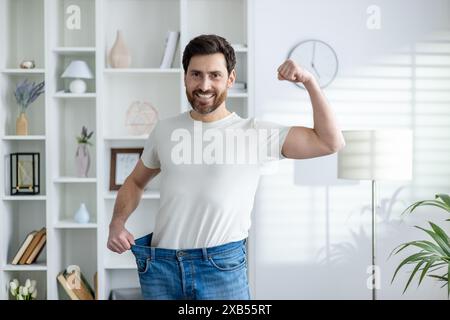 Portrait of a happy and confident young man at home, showing his weight and waist in oversize jeans, showing a sit and victory gesture with his hand. Stock Photo