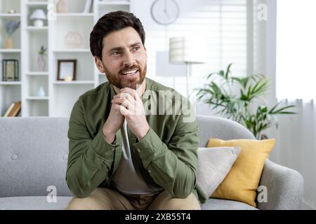 A smiling young man is sitting on the sofa at home, holding his hands together near his face and looking to the side with satisfaction. Stock Photo