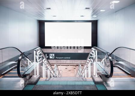 Two escalators and staircase leading down to a passport control area with a large blank billboard above for advertisement mockup. The scene is set in Stock Photo