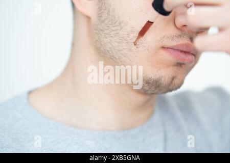young man applying beard growth oil with pipette close up. man holding pipette with oil for beard, applying cosmetic serum with pipette onto face Stock Photo