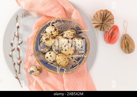 Quail eggs in a ceramic bowl with a peach fuzz pink colored scarf and bird feathers. Wooden table painted white. Background with elements suggestive o Stock Photo