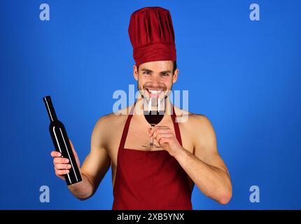 Professional male sommelier tasting and sniffing red wine in wineglass. Winemaking. Happy winemaker in burgundy cook hat and apron with bottle of wine Stock Photo