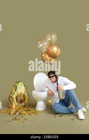 Young man with hangover and champagne sitting near toilet bowl after Birthday party on green background Stock Photo