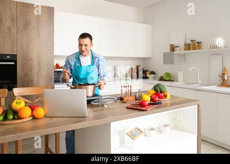 Young man with laptop cooking soup in kitchen Stock Photo