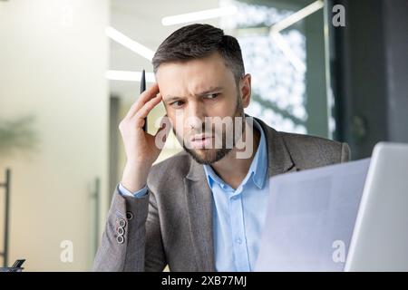 Businessman in office looking confused while reading a document and working on a laptop, holding a pen in hand. Stock Photo