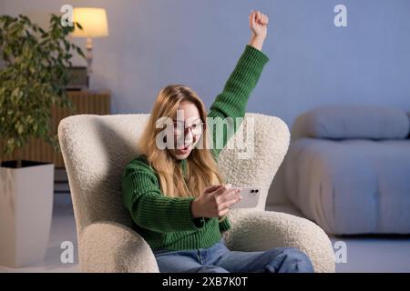A woman is comfortably seated in a chair, holding a cell phone with her arms raised. The room has a couch, plants, and a joyful atmosphere Stock Photo
