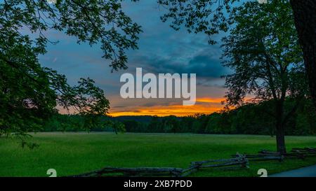 A sunset over a grassy field with trees and a fence beside the street Stock Photo