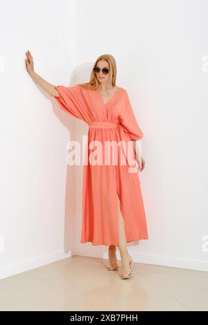 A stylish blonde woman in a coral maxi dress with a v-neckline leaning against white wall Stock Photo