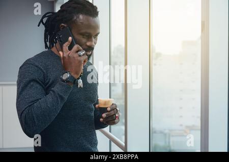 African adult man with dreadlocks on a phone call listening while looking through the window. is in the office, standing, smiling and he is enjoying h Stock Photo