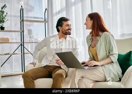 A redhead woman and bearded man are seated together, focusing on a laptop screen as they spend quality time at home. Stock Photo