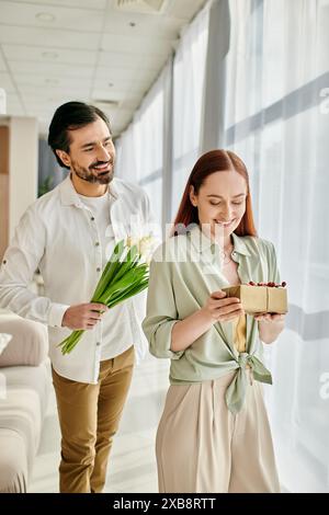A bearded man and a redhead woman are walking through a modern living room, holding a gift box. Stock Photo